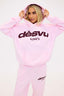 Person wearing a pink tracksuit with burgundy dés vu logo on a white background