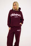 Person wearing a burgundy tracksuit with  dés vu logo in pink on a white background