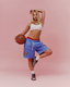 A blonde woman poses in a white sports bra and blue icon mesh shorts, holding a basketball against a solid pink studio background.