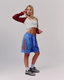 A woman modeling blue icon mesh shorts with a large orange 'dès vu' logo, paired with a white and maroon cropped long-sleeve top, white socks, and black-and-white sneakers against a white studio background.