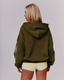 A woman shown from the back wearing an oversized olive green hoodie with a yellow graphic print on the left sleeve, paired with light cream shorts, against a solid gray studio background.