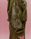 An olive green sweatshirt with large yellow abstract line-art embroidery on the sleeve, shown on a model against a solid pink background.