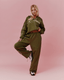 A blonde model poses in a matching olive green cropped hoodie and sweatpants set with yellow graphic line details, set against a solid pink studio background.