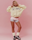 A model posing in a pastel yellow zip-up hoodie with a navy script logo, paired with a pink wrap skirt, gray athletic shorts, cream leg warmers, and black-and-white sneakers, set against a solid pink studio background.