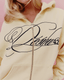 A close-up of a person wearing a yellow colored zip-up hoodie with a large navy cursive script graphic across the chest. The background is a solid light pink.