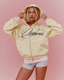 A woman wearing a pale yellow zip-up hoodie with a navy script logo on the chest, paired with light pink and gray athletic shorts, posing against a solid pink background.