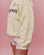 A person wearing a yellow colored oversized hoodie with navy graphic text on the back and raised textured embroidery on the sleeve, paired with gray shorts, against a solid pink background.
