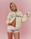 A blonde model wearing a yellow colored zip-up wiith a navy script logo that reads 'desvu 422', paired with light pink and gray athletic shorts, holding a green drink against a solid pink background.