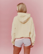 A woman shown from behind wearing a pale yellow cropped hoodie and light gray heathered athletic shorts against a solid pink studio background.