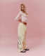 A woman posing in a light pink long-sleeve crop top and yellow colored wide-leg sweatpants with navy graphic script on the side, set against a solid pink studio background.